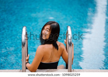 A young Chinese Asian woman lounges by the side of a pool by the ladder. She is young, tanned and athletic and is smiling at the camera. 