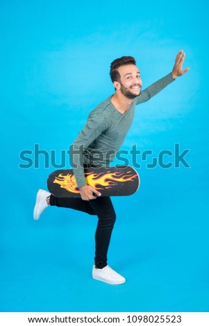 Full-length shot of young man holding the skateboard dancing raising hand and leg up back on a blue background.