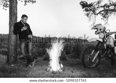 motorcyclist man is stands by the fire holds a metal cup and uses a smartphone. camping in the mountains forest. tourist have a rest at a campfire near tent. off road motorcycle adventure, enduro, bw