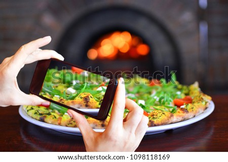 Girl takes photo of pizza on smartphone camera. Pizza in front of a stone stove with fire. Background for a traditional pizzeria restaurant. 