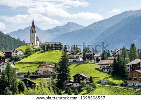 Summer scenery of beautiful Schmitten village in Graubünden, Switzerland, with view of houses on green grassy hills, a lovely church on hilltop & majestic mountains in background under clear blue sky