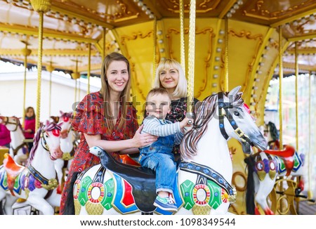 Cute little boy sitting on carousel horse while his mother with the girlfriend standing next to them and holding them slightly.