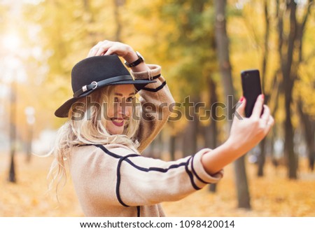 Young pretty woman in black hat making selfie with her smartphone in the autumn park full of yellow leaves