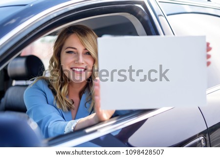Woman sitting in the car and holding a white blank poster. Attractive blonde with a clean sheet of paper or your text. Focus on model.