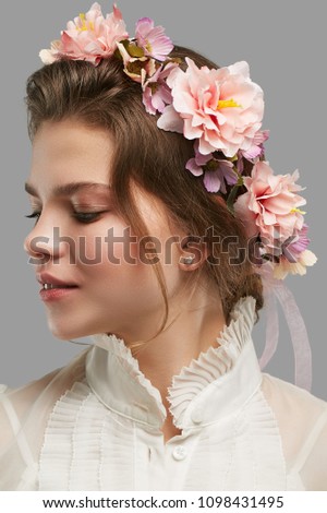 Profile portrait of a young lady with a bohemian flower crown, in a white vintage blouse. The attractive girl looking down pensively on the grey background, light pink peonies and daisies in her hair.