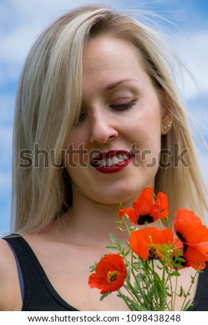 European caucasian happy young woman, blonde with open shoulders in a black dress with red lips and a snow-white smile costs in the blossoming field with a bouquet of red poppies under the blue sky.