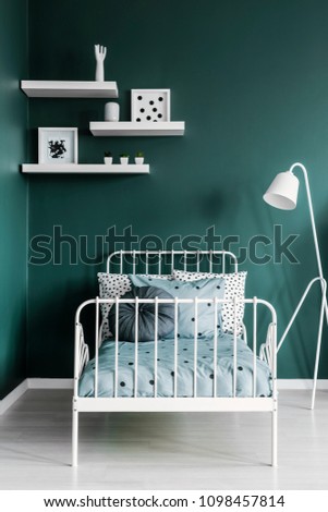Shelves with decorations and a floor lamp above a white, metal frame twin bed in a dark green room interior for a teenager