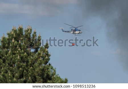 Fire rescue police helikopter delivering water bucket for aerial firefighting, smoke and blurred pine tree, blue sky byckground