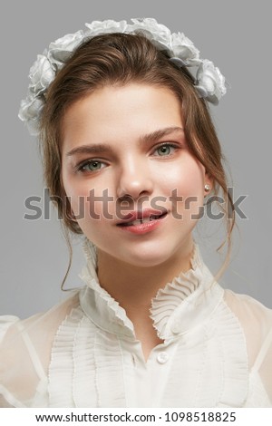 Portrait of a young lady with a flower crown, in a white vintage blouse. The girl looking pensively at the camera over the grey background, lips parted into a tender smile, white roses in her hair.