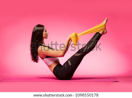 Long haired beautiful pilates or yoga athlete does a graceful pose with a yellow rubber band while wearing a tight sports outfit against a pink background in a studio Royalty-Free Stock Photo #1098550178