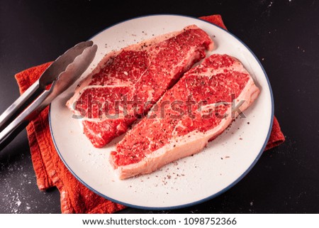 Two cuts of strip steak resting on a plate being prepared with salt and pepper for cooking.
