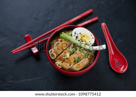Ramen noodles with roasted chicken meat and egg served in a red tableware, elevated view over black stone background