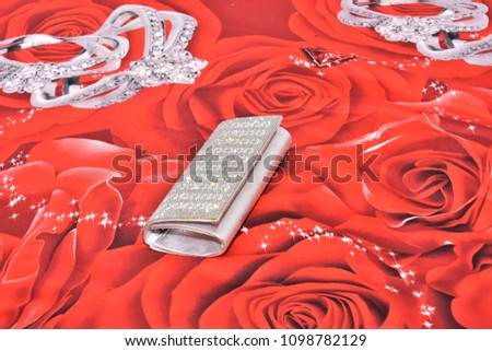 Silvery small female handbag clutch on scarlet quilt with pattern of large red roses and decorations on the bed
