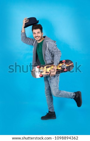 Full-length shot of a handsome young man wearing a casual outfit standing, holding his skateboard and his hat, looking clumsy, standing on a blue background