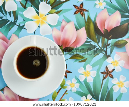 White cup of coffee with a white saucer on a blue background with a picture of flowers and green leaves, top view