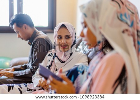 Two young Muslim women chat while sitting on the couch during Ramadan. They are talking animatedly and are both wearing hijab headscarves.