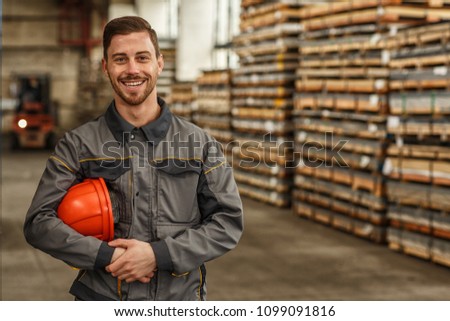Attractive young male constructionist smiling to the camera, holding his hardhat, posing at the storage of metalworking factory. Profession, heavy industry, achievement, development concept Royalty-Free Stock Photo #1099091816