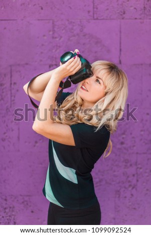 Portrait of beautiful smiling blonde girl, female, woman in black t-shirt and pants, making photos at summer street. Professional photographer in casual, holding photo camera. Background purple color.