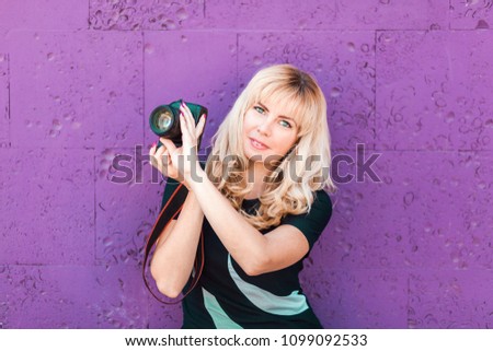 Portrait of beautiful smiling blonde girl, female, woman in black t-shirt and pants, making photos at summer street. Professional photographer in casual, holding photo camera. Background purple color.