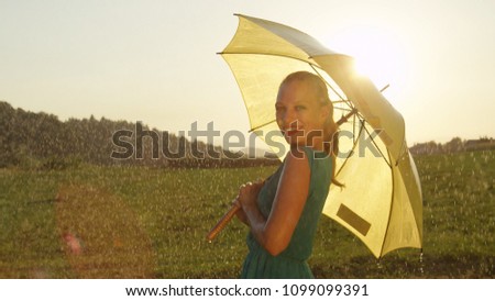 PORTRAIT LENS FLARE: Gorgeous blonde woman in a green sundress smiles over her shoulder while twirling in the picturesque meadow under yellow umbrella. Beautiful golden sun rays shine on woman dancing