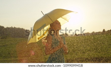 SUN FLARE, CLOSE UP: Smiling blonde woman looks around the picturesque rural landscape during a refreshing summer shower. Carefree girl enjoying the sights as while she spins under yellow umbrella.