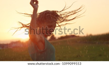 CLOSE UP, LENS FLARE: Joyful blonde girl enjoys her evening in the countryside by dancing in the rain. Stunning golden sun rays shine on playful young woman spinning and enjoying a spring shower. Royalty-Free Stock Photo #1099099553