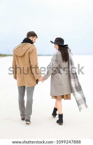 Charming woman and man walking on snow and holding hands, wearing coat, grey scarf and hat. Concept of seasonal inspiration and couple photo session.