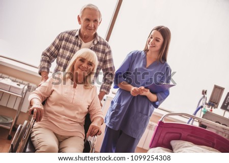 Beautiful old lady is sitting in the wheelchair in hospital ward, her husband and a nurse are near, all are smiling