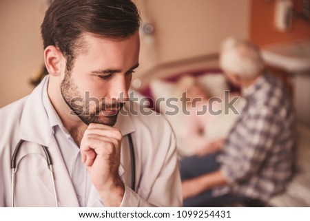 Old man is making a visit to his wife who is lying in bed in the hospital ward, handsome doctor in the foreground