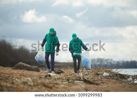 Two men with big sacks walking along river bank, picking up litter and putting it in those sacks Royalty-Free Stock Photo #1099299983