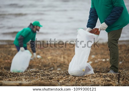 One of two greenpeacers putting smashed plastic bottle into big litter sack while cleaning territory of river bank Royalty-Free Stock Photo #1099300121