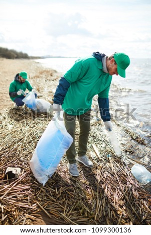 Young greenpeacers in uniform walking along waterside and picking up litter for utilization Royalty-Free Stock Photo #1099300136
