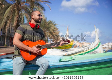 Handsome musician wearing a casual outfit, sitting on a boat and holding his guitar playing music.