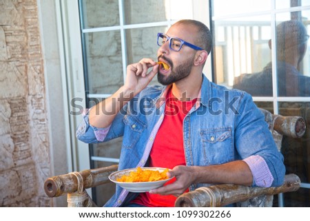 Close shot of a handsome man wearing a casual outfit, sitting in the home and holding a plate full of food and eating it, looks that he is watching tv.