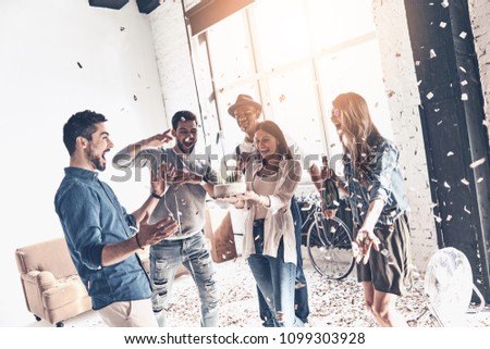 Creating happy memories. Happy young man celebrating birthday among friends while standing in room with confetti flying around Royalty-Free Stock Photo #1099303928