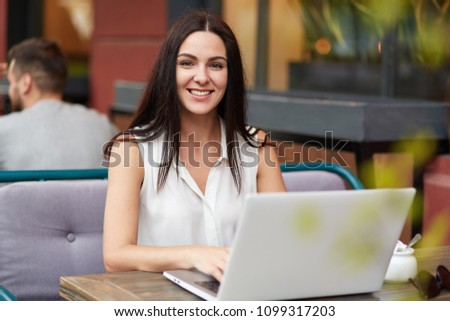 Successful female freelancer works remotely on laptop computer, sits in outdoor cafe, has pleasant smile, enjoys remote work, looks happily at camera. People, leisure and technology concept.