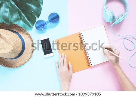 Creative flat lay woman's hands with notebook and smartphone, hat, sunglasses and headphones on pastel color background, Summer vacation concept