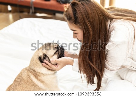 Beautiful Asian young woman playing with her dog and smile with dog pug breed looking in funny face in bedroom,Friendship Concept