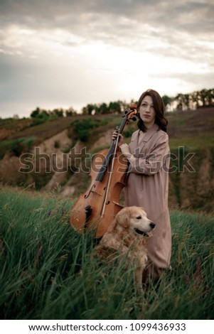 Woman cellist prepares to play at meadow against backdrop of picturesque landscape next to dog.
