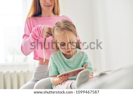 Young mother styling daughter's hair while she's watching cartoons on smart phone