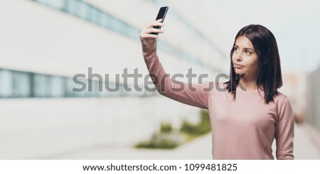 Young pretty woman confident and cheerful, taking a selfie, looking at the mobile with a funny and carefree gesture, surfing the social networks and internet in the university campus