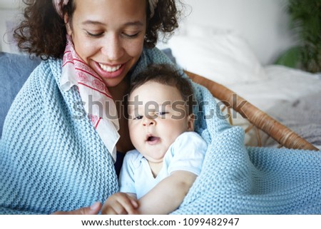 Portrait of cute sleepy baby boy yawning sitting on mother's lap in cozy light bedroom. Happy smiling young mixed race female spending lazy morning at home, hugging her adorable toddler son