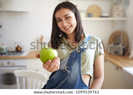 Indoor shot of cheerful stylish girl in her twenties choosing healthy lifestyle, posing in kitchen, handing over juicy organic green apple at camera and smiling. Selective focus on woman's hand