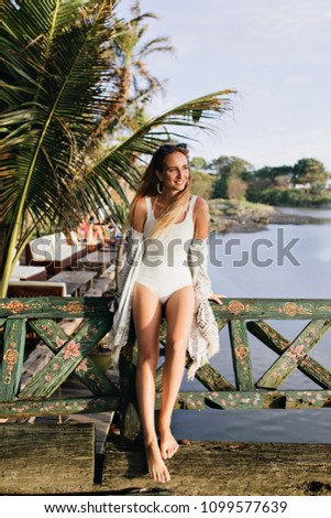 Full-length portrait of blissful slim girl looking away during photoshoot in exotic place. Joyful tanned lady standing near palm trees and smiling.
