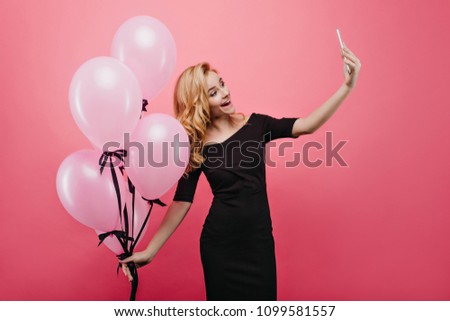 Shapely fascinating young woman using new phone for selfie. Laughing birthday girl taking picture of herself while holding big bunch of party balloons.