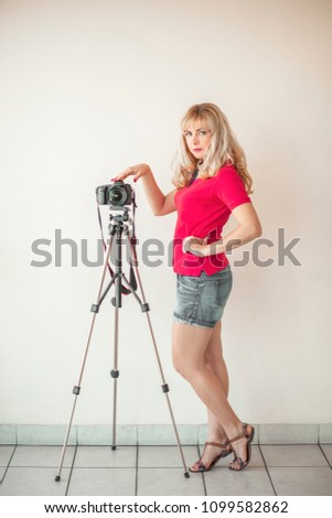 Full-length portrait of beautiful smiling blonde girl, female, woman in red t-shirt, making photos at summer street. Portrait of professional photographer in casual wear holding photo camera at tripod