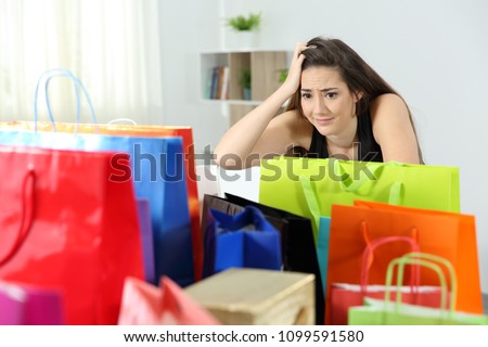 Worried shopaholic woman after multiple purchases in colorful shopping bags at home Royalty-Free Stock Photo #1099591580