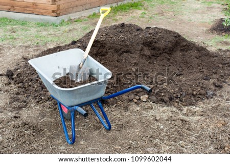 The wheelbarrow stands near a large pile of humus.  In the wheelbarrow is a shovel. In the background a fence of boards. There is no one in the picture.