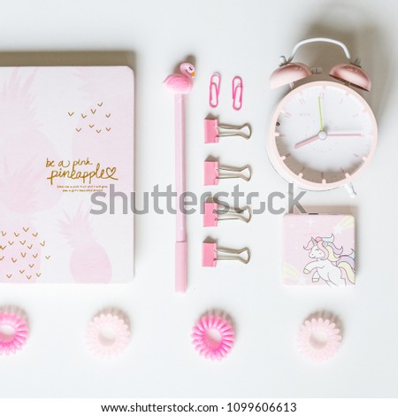Desktop flatlay pink color: notebook with pineapples, pen with flamingo, clips for paper, alarm clock, hair bands and mirror with unicorn lying on white background