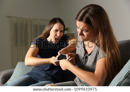 Two friends or sisters fighting for a smart phone sitting on a couch in the living room at home Royalty-Free Stock Photo #1099610975